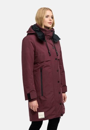 Woman wearing a long maroon winter coat with black zippered pockets and a high collar with drawstrings, standing against a plain background.