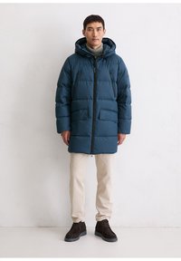 Doudoune bleu marine à capuche, avec une fermeture éclair, deux poches avant et une coupe ample, portée par-dessus un pull léger et un pantalon beige.