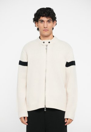 Cream knitted sweater with a high collar, front zipper, and black stripe detail on the sleeves. Soft texture and relaxed fit.