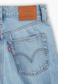 Poche arrière de jean en denim bleu clair avec une étiquette en cuir marron Levi Strauss & Co. et une étiquette en tissu rouge Levi's sur la couture de la poche.