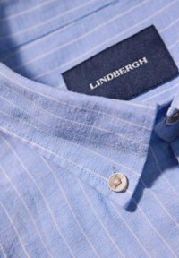 Lichtblauw overhemd met kraag en witte strepen; heeft een knoopsluiting en een marineblauw label met de tekst "LINDBERGH." Gestructureerde stof.