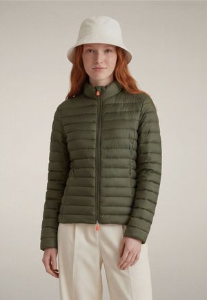 Woman with red hair wears white bucket hat, olive green quilted jacket zippered up, and cream-colored pants, standing against plain background.