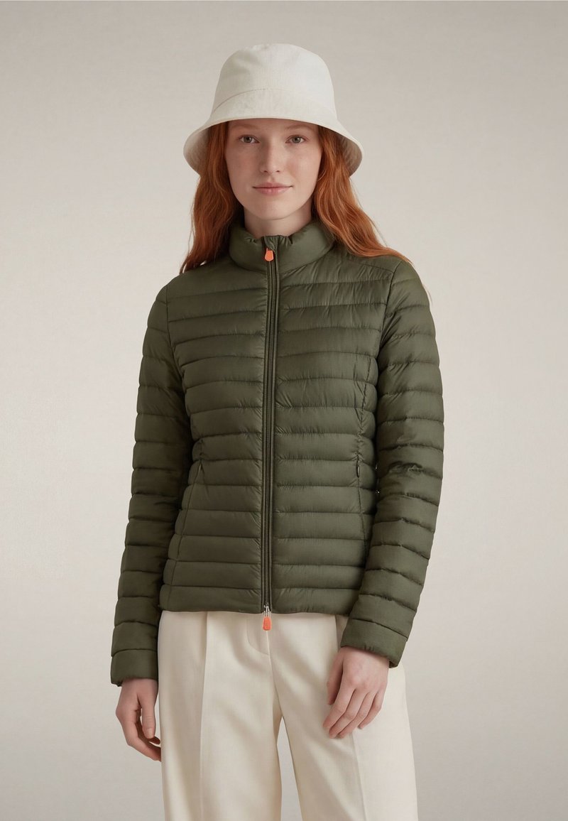 Frau mit roten Haaren trägt weißen Fischerhut, olivgrüne gesteppte Jacke mit Reißverschluss und cremefarbene Hose, steht vor einfachem Hintergrund.