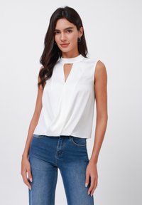 White sleeveless top with a high neckline and keyhole detail, featuring a draped front and a textured finish. Paired with blue denim jeans.
