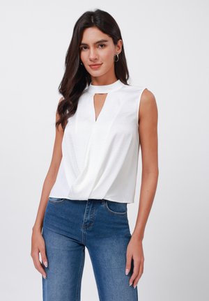 Hvit ærmeløs topp med høy hals og nøkkelhulldetalj, med en drapert front og teksturert finish. Kombinert med blå denimbukser.