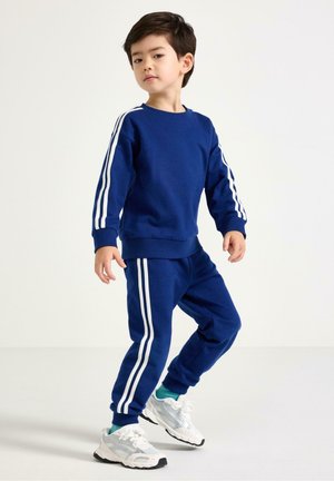 Nuori poika seisoo, päällään sininen verkkatakki valkoisin raidoin ja valkoiset tennarit, yksinkertaista valkoista taustaa vasten.