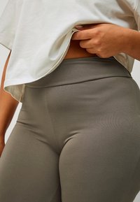 Persoon heft witte shirt op om high-waisted, gladde grijze legging op heupen en bovenbenen te onthullen.