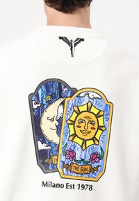 Hvit hettegenser med fargerike grafiske trykk av solen og månen, omrisset i svart med blomsteraksenter. Teksten sier "Milano Est 1978."