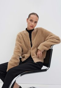 Giacca in pile beige con zip frontale, polsini e orlo a costine. Il modello è seduto su una sedia nera, indossando una maglia nera e pantaloni neri.