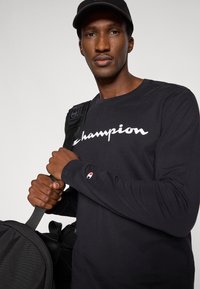 Maglia nera a maniche lunghe con logo "Champion" bianco, polsini a coste e emblema rosso sulla manica. Il modello tiene una borsa nera.