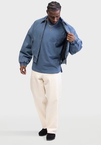 Denim bomber jacket, blue T-shirt, cream pants, and black shoes. Jacket has elastic cuffs and a zip front; T-shirt features a chest pocket.