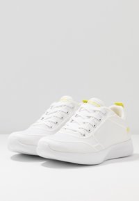 Baskets de sport blanches avec un matériau en maille, un design à lacets, une semelle texturée et de subtils accents jaunes au niveau du talon et du branding.