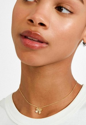 Gouden ketting met een fijne ketting en een bloemvormige hanger met drie heldere edelstenen. Draagt bij een lichtgekleurd shirt.