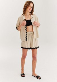 Béžová lněná košile s černým lemováním ve tvaru vlny, spárovaná s odpovídajícími šortkami s podobným lemem. Černé sandály dotvářejí vzhled.