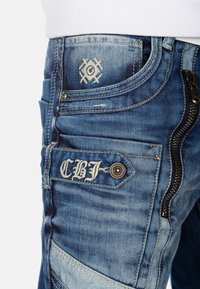 Jeans in denim blu chiaro con cuciture a contrasto, tasca con zip, dettagli ricamati e cuciture decorative sulla parte anteriore.