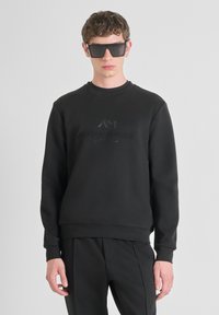 Felpa in cotone nera con collo a giro e logo in rilievo sul petto, abbinata a pantaloni neri coordinati. Texture liscia, design minimale.