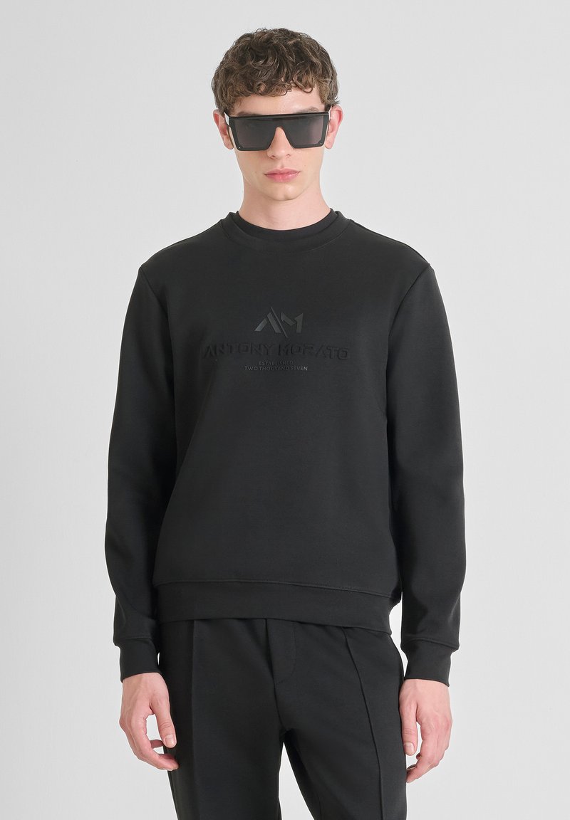 Felpa in cotone nera con collo a giro e logo in rilievo sul petto, abbinata a pantaloni neri coordinati. Texture liscia, design minimale.