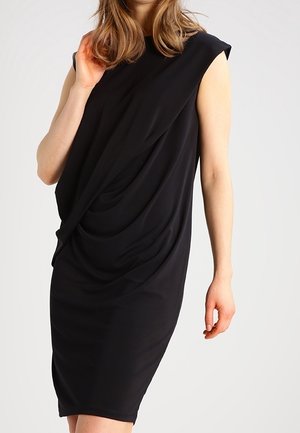 Mujer que lleva un vestido negro sin mangas con detalle de tela drapeada en la parte delantera, de pie frente a un fondo claro y liso.