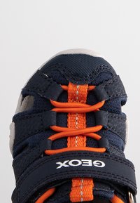 Scarpe sportive blu navy e grigie con lacci arancioni, tomaia in mesh per traspirabilità e suola in gomma. Logo Geox sulla fascia laterale.
