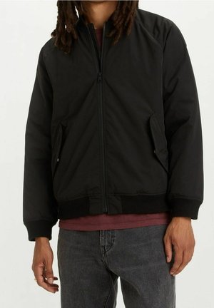 Osoba ubrana w czarną kurtkę na zamek błyskawiczny, pod którą ma bordową koszulę, oraz szare dżinsy, stojąca z rękami swobodnie opuszczonymi wzdłuż ciała.