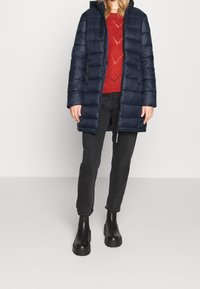 Marinblå vadderad lång kappa med huva, bärs över en röd stickad tröja. Matchad med svarta jeans och kraftiga svarta ank stövlar.