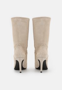 Botas de tacón alto de ante beige con un caño ajustado, punteras puntiagudas y tacones de aguja. Textura suave con un detalle de costura a lo largo de la parte posterior.