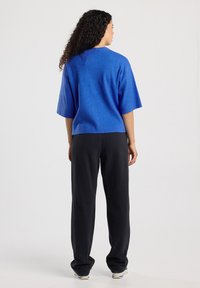 Vrouw met krullend haar staat met haar rug naar ons gekeerd, draagt een blauwe korte mouwtop, zwarte losse broek en witte sneakers op een effen achtergrond.