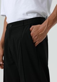 Zwarte broek met een elastische tailleband, voorzien van een enkele knoopsluiting, zijzakken en een gladde, strakke textuur.