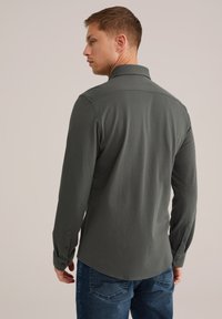 Camisa de manga larga gris oscuro con cuello de punta, puños abotonados y dobladillo curvado. La tela parece suave, ajustándose de manera holgada al modelo.