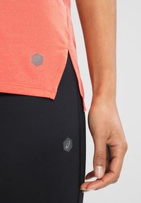 Top sportivo corallo con finitura texture, spacco laterale e logo esagonale. Leggings neri con superficie liscia e dettaglio logo discreto.