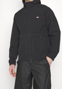 Svart hooded jacka med elastiska ärmslut, dragkedja framtill och fickor. Har kontrasterande sömmar och en logotyp på bröstet.