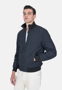 Giacca bomber navy con colletto alto, zip frontale e tasche laterali. Presenta polsini e orlo a coste neri con una texture di tessuto liscia.