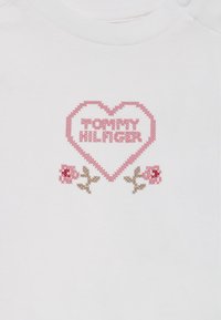 Vit bomull t-shirt med en rosa hjärt-grafik som har texten "TOMMY HILFIGER" och blomsterdetaljer i rosa och brunt.