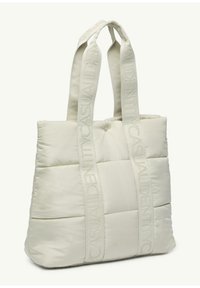 Cream-colored quilted tote bag with textured fabric, featuring padded sides and vertical logo straps for added detail.