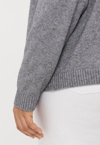 Harmaa neulepaita, jossa on ribbireunus, kuvattu sivusta. Materiaalin näyttäisi olevan pehmeää ja teksturoitua, yhdistettynä valkoisiin housuihin vyötäröllä.