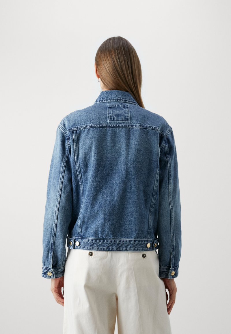 Persona con cabello largo y castaño, vistiendo una chaqueta de denim azul y pantalones blancos, de espaldas frente a un fondo blanco liso.