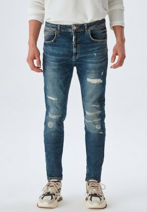 Blue denim jeans with a slim fit, featuring distressed areas and light fading. Worn with beige and black patterned sneakers.