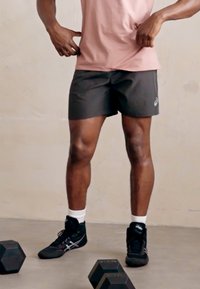 Sorte træningsshorts med et logo, en tætsiddende pink t-shirt og sorte sneakers med hvide detaljer; står med vægte på en grå overflade.