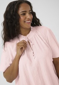 Haut rose à manches courtes en tissu texturé, avec un col à volants et un détail noué au col ; présente des accents de boutons à l'avant.