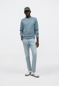 Ανοιχτό μπλε βαμβακερό φούτερ με στρογγυλή λαιμόκοψη, σε συνδυασμό με ανοιχτόχρωμο τζιν και αθλητικά παπούτσια σε μαύρο και άσπρο. Το γκρι καπέλο διαθέτει υφασμένο λογότυπο.