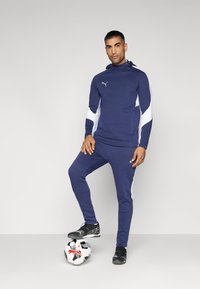 Marineblauwe sporttracksuit met witte zijaccenten. Voorzien van een kap en elastische manchetten. Zwarte sportschoenen met een wit en zwart voetbal.