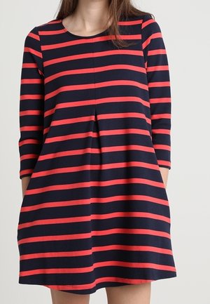 Femme portant une robe bleu marine à rayures rouges horizontales et manches trois-quarts, debout devant un fond clair uni.
