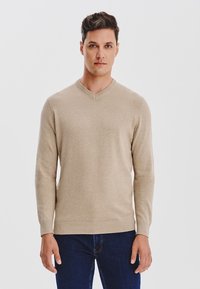 Beżowy sweter w serek wykonany z miękkiego materiału, z długimi rękawami i prążkowanym wykończeniem, w zestawie z ciemnymi dżinsami.