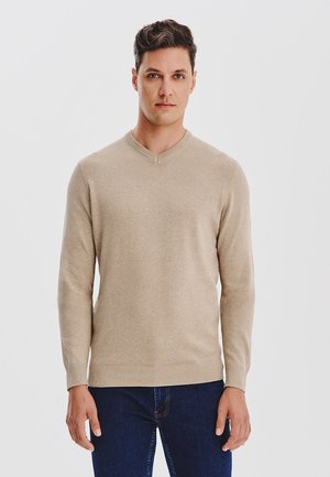 Beżowy sweter w serek wykonany z miękkiego materiału, z długimi rękawami i prążkowanym wykończeniem, w zestawie z ciemnymi dżinsami.