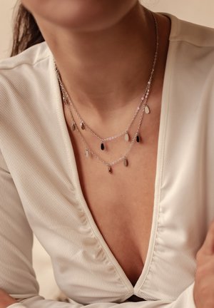 Dreischichtige Silberkette mit mehreren Tropfen-Anhängern in Schwarz, Weiß und metallischen Oberflächen, die an filigranen Ketten hängen.