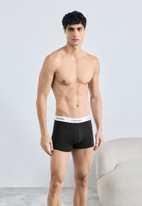 Czarne bokserki z geometrycznym wzorem, biały elastyczny pasek z logo Calvin Klein, model stoi na neutralnym tle.