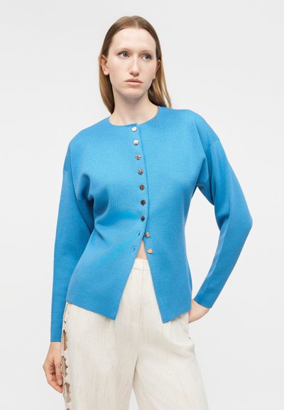 Γυναίκα με μπλε ζακέτα με κουμπιά και λευκό παντελόνι με δαντελένια λεπτομέρεια στο πλάι, στέκεται με το ένα χέρι στη μέση μπροστά από απλό φόντο.