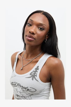 Hvid tanktop med grå grafiske designs, lagdelte sølv chunky kæder og hoop øreringe; glat hud, langt glat sort hår.