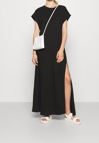 Černé maxi šaty s krátkým rukávem, bočním rozparkem, v kombinaci s bílou kabelkou přes rameno a bílými sandály na pásky. Hladká textilie.