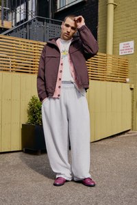 Giacca trapuntata bordeaux con interno rosa, t-shirt bianca, pantaloni di felpa grigi e scarpe slip-on viola. In piedi all'aperto contro uno sfondo di legno.